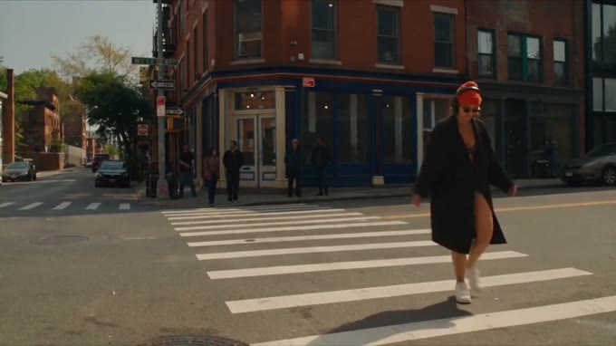 Wide street-level shot: the lead in a red cap and headphones crosses at the lights.